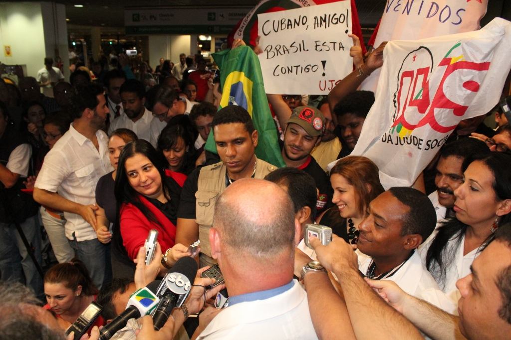 Cuban doctors surrounded by reporters and supporters holding banners at an airport arrivals hall in Brasília