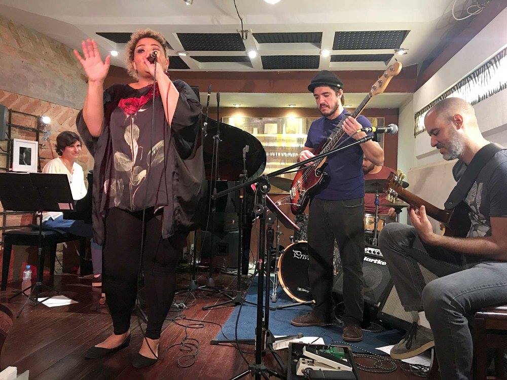 A woman singing into a microphone with a small band behind her in an intimate Rio de Janeiro music room