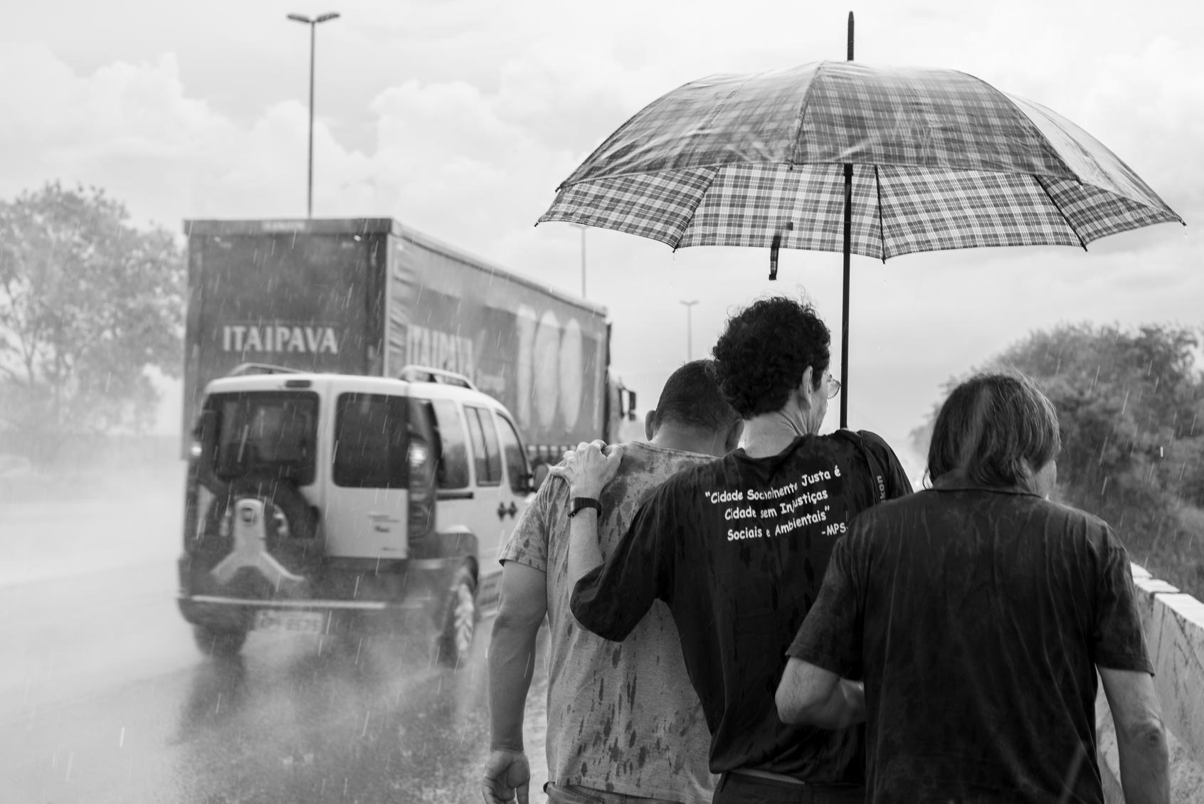 Three man walking over highway bridge in heavy rain sharing one umbrella. Image shot 01/2018. Exact date unknown.
