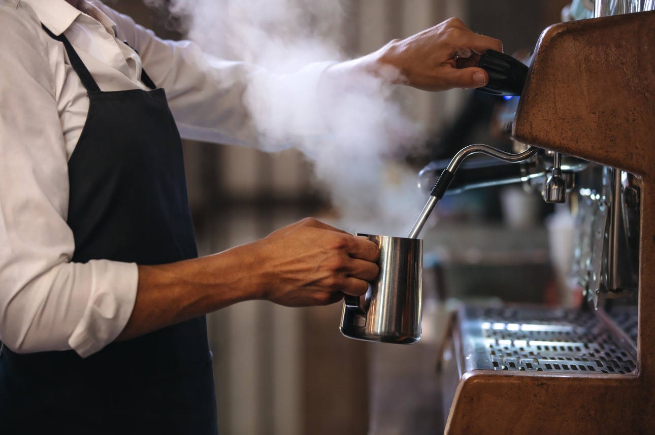 Coffee shop worker preparing coffee on steam espresso coffee machine. Cropped shot of man working in coffee shop wearing an apron.