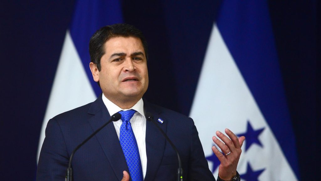 Man in suit speaking at podium with Honduran flags