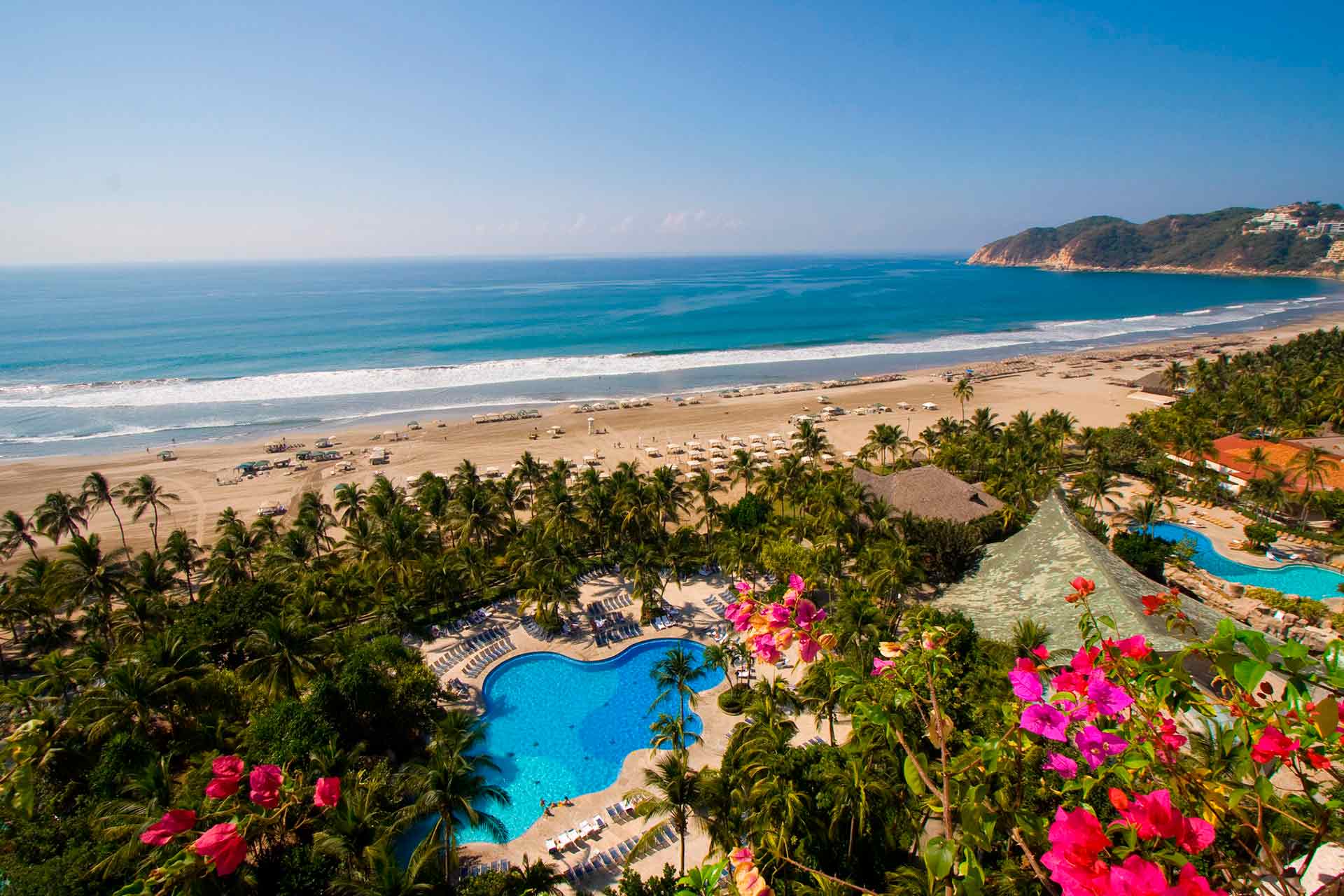 Aerial view of a palm-lined beach resort with swimming pools on a Pacific bay
