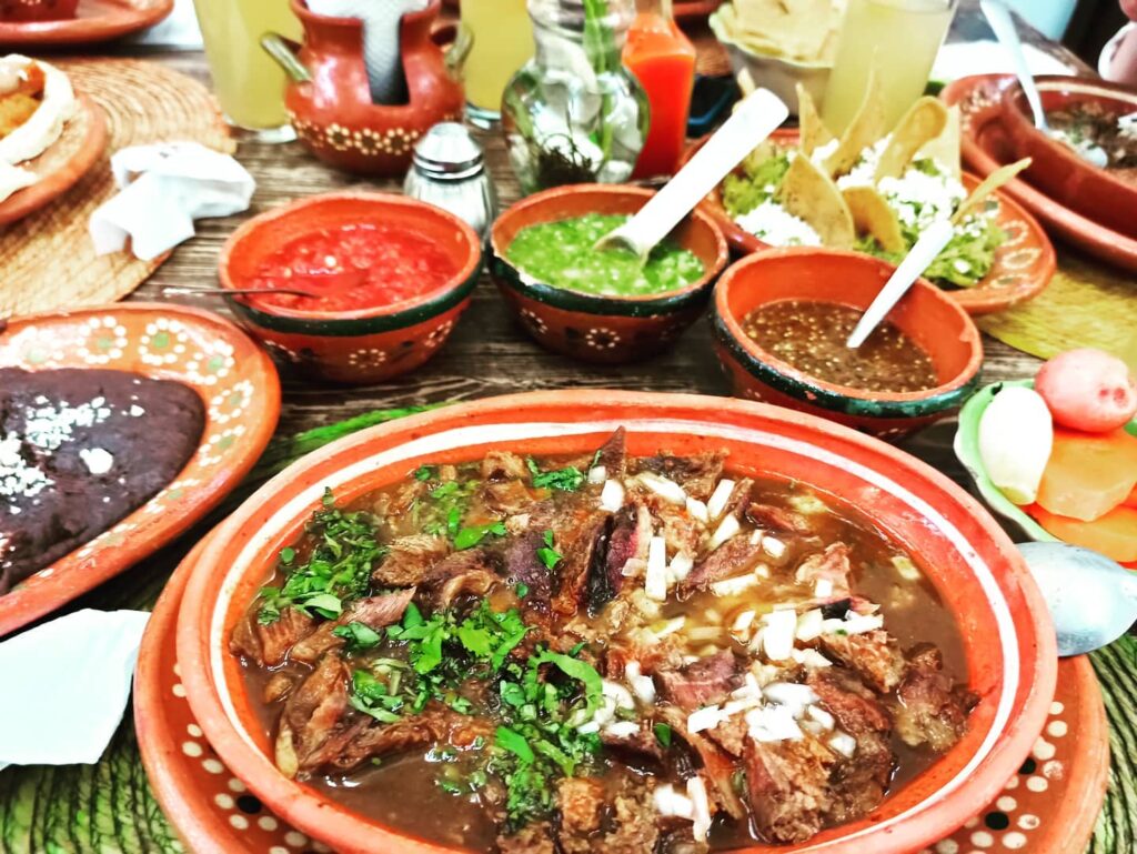 Traditional birria de chivo goat stew served in a clay dish with salsas