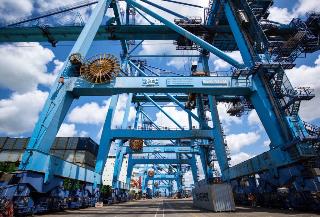 Large blue port cranes and shipping containers under a cloudy sky