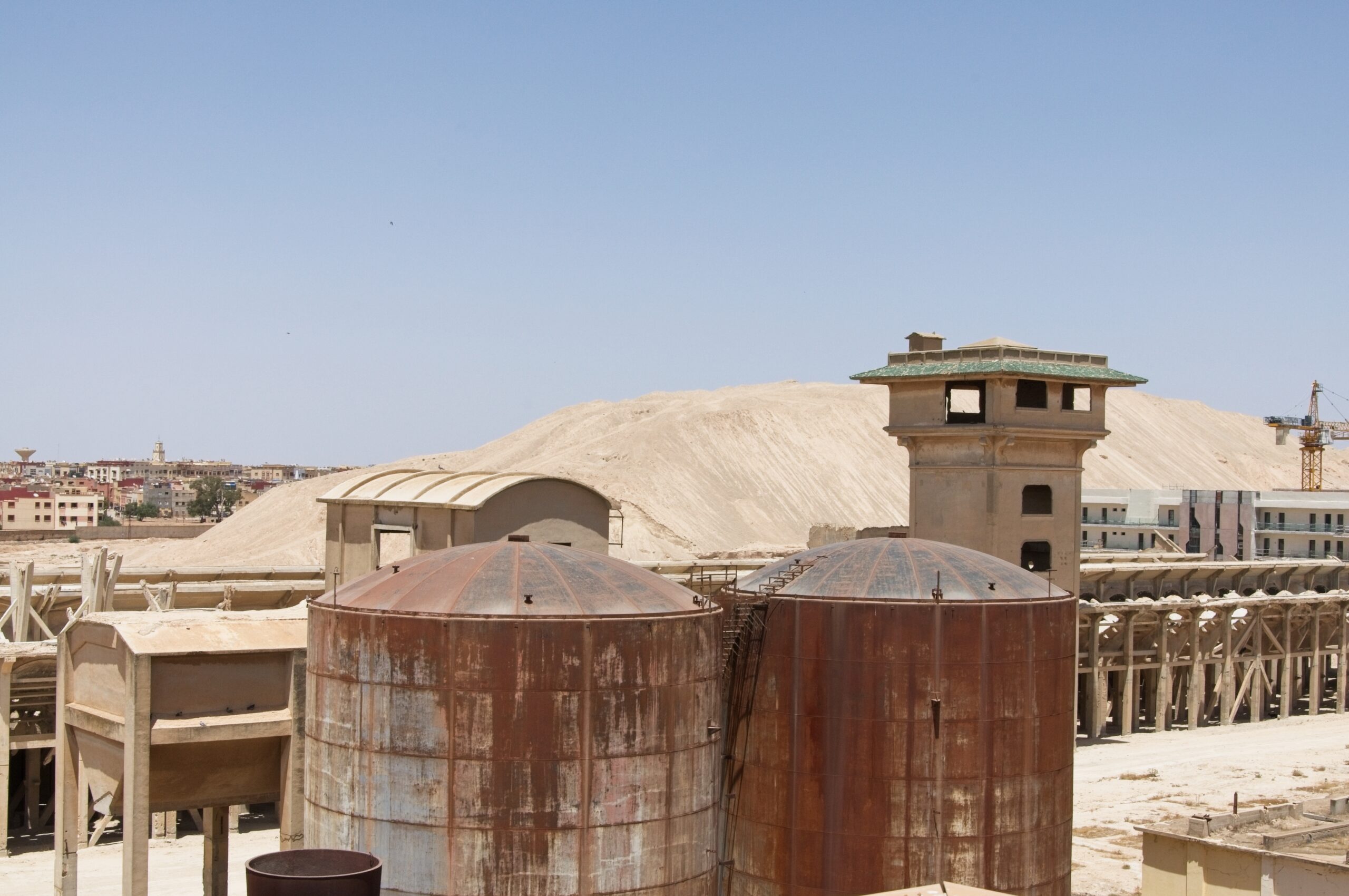 Rusted storage tanks and a concrete tower at an old phosphate works at Khouribga, with a pale spoil heap and the town behind.