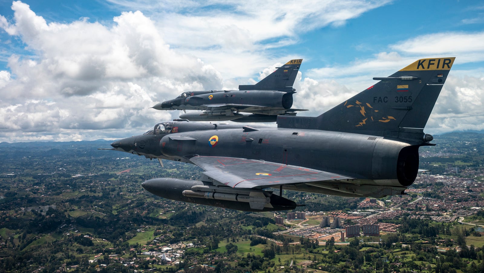 Two Colombian Air Force Kfir fighter jets flying in formation over green hills and a town