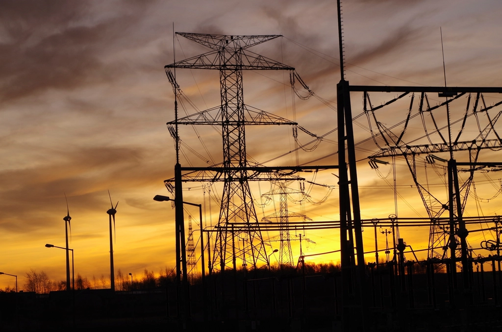 Silhouetted high-voltage transmission towers and wind turbines against an orange sunset sky