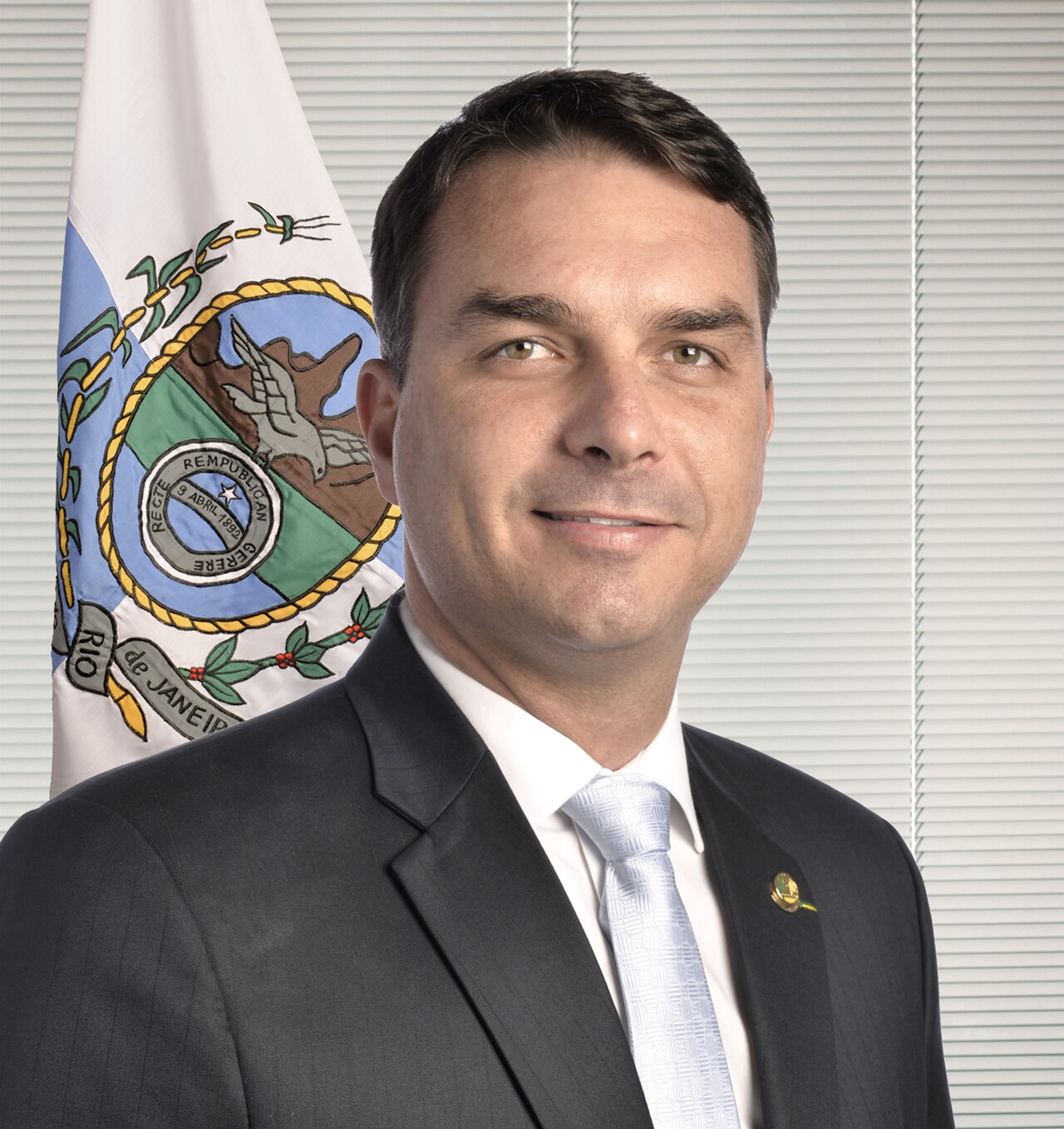 Senator Flávio Bolsonaro in a dark suit beside the flag of the state of Rio de Janeiro