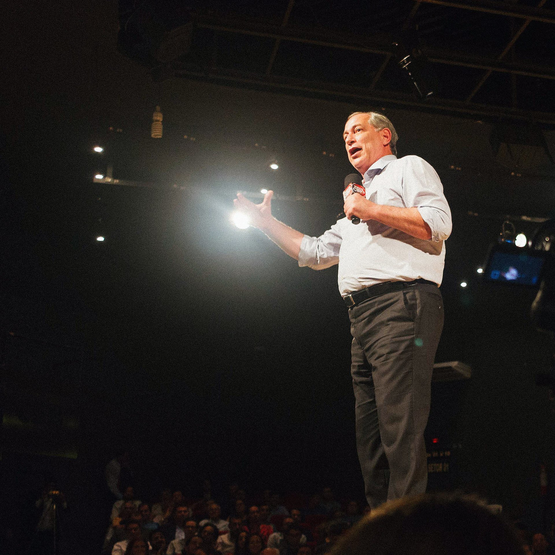 Ciro Gomes speaks into a microphone on stage at a public event, addressing a seated audience.
