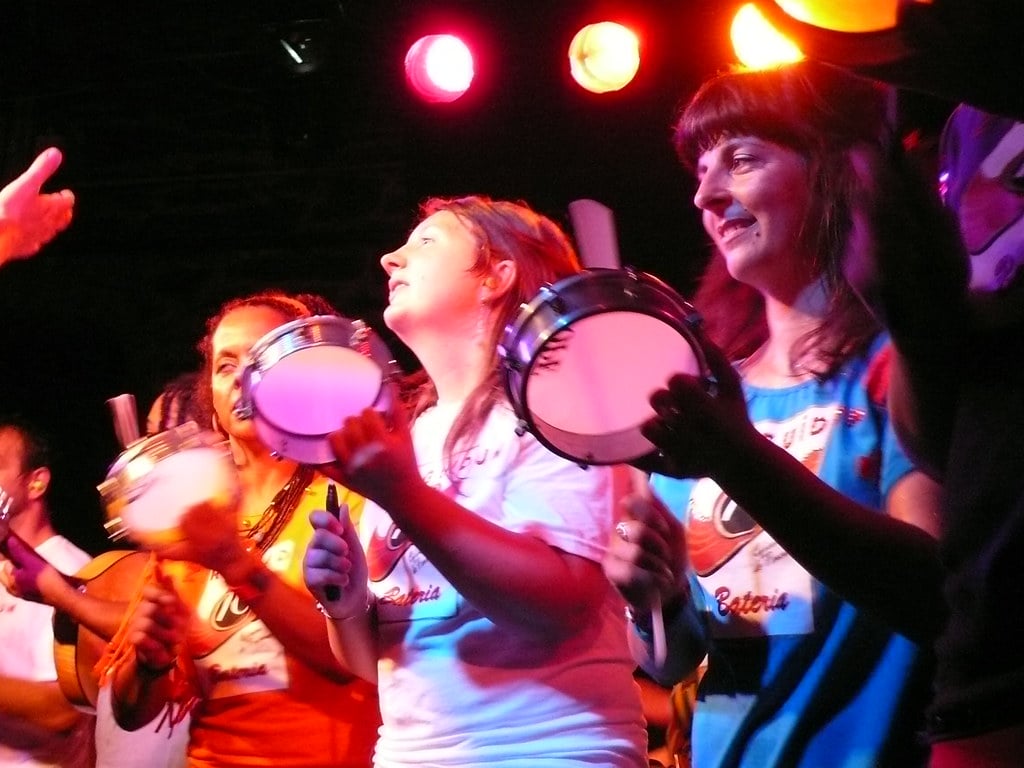 Musicians playing tamborim frame drums under stage lights at a samba performance