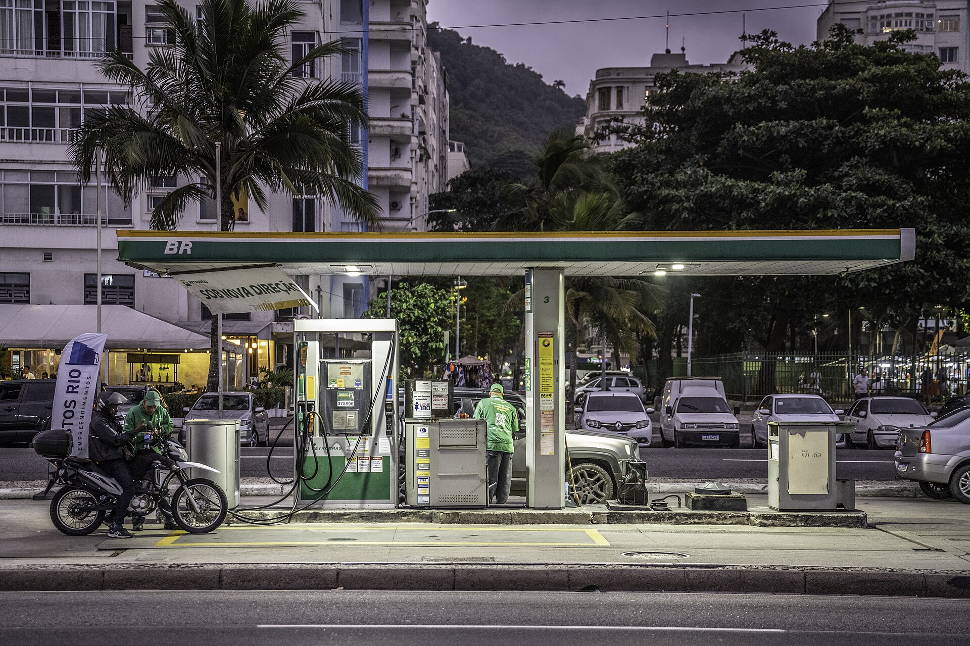Attendant at a BR-branded filling station on a Rio de Janeiro avenue at dusk, with a motorcyclist at the pump and apartment blocks behind
