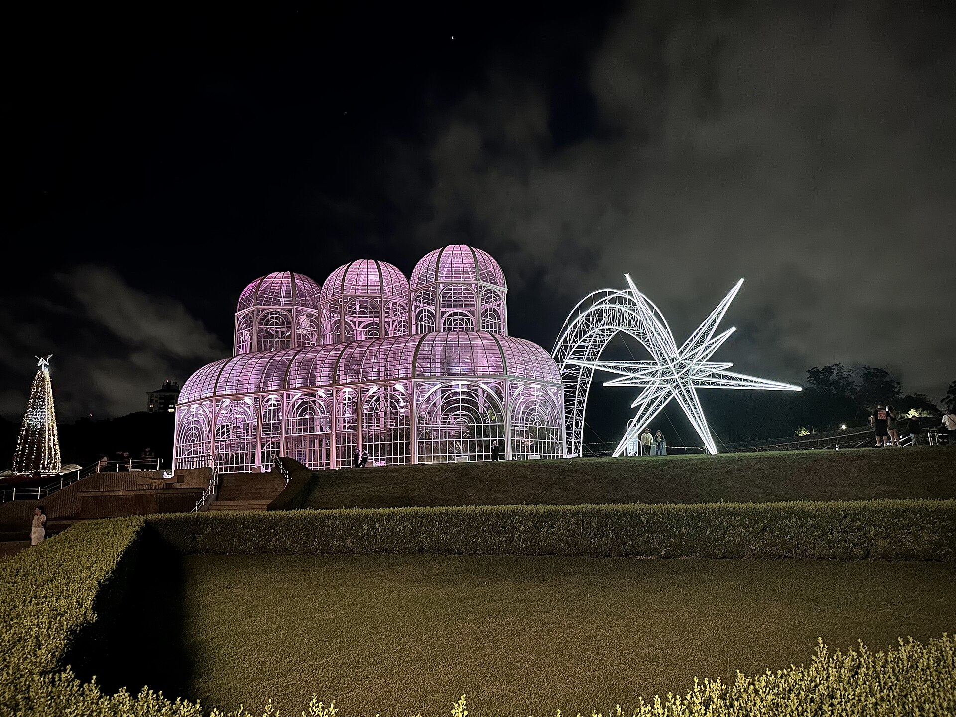 The glass and iron greenhouse of Curitiba's botanical garden lit pink at night beside an illuminated star sculpture.