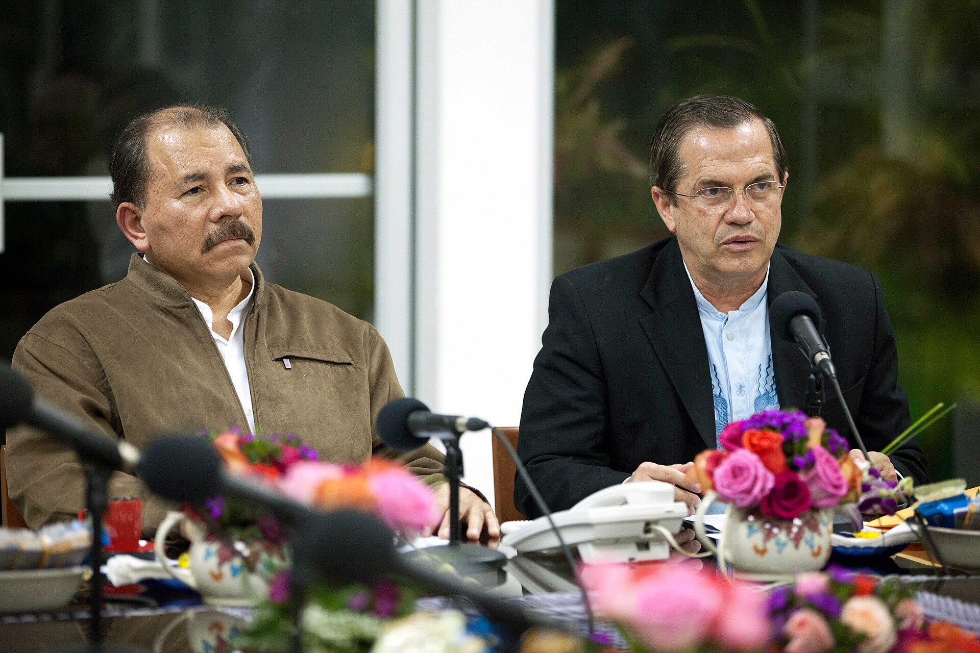 Daniel Ortega, seated at a table with microphones and flowers, beside a man in a dark suit speaking
