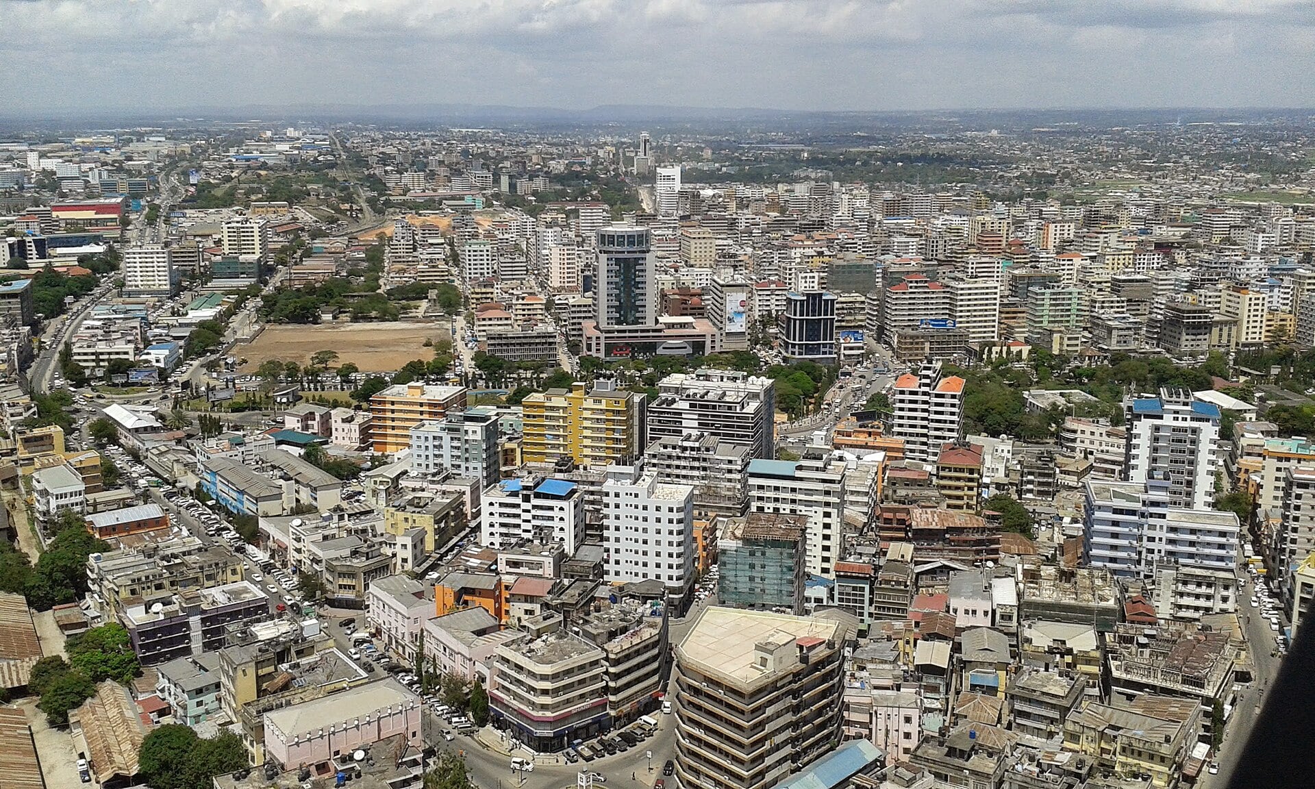 An aerial view across Dar es Salaam showing dense mid-rise buildings, main roads and distant hills under cloud.