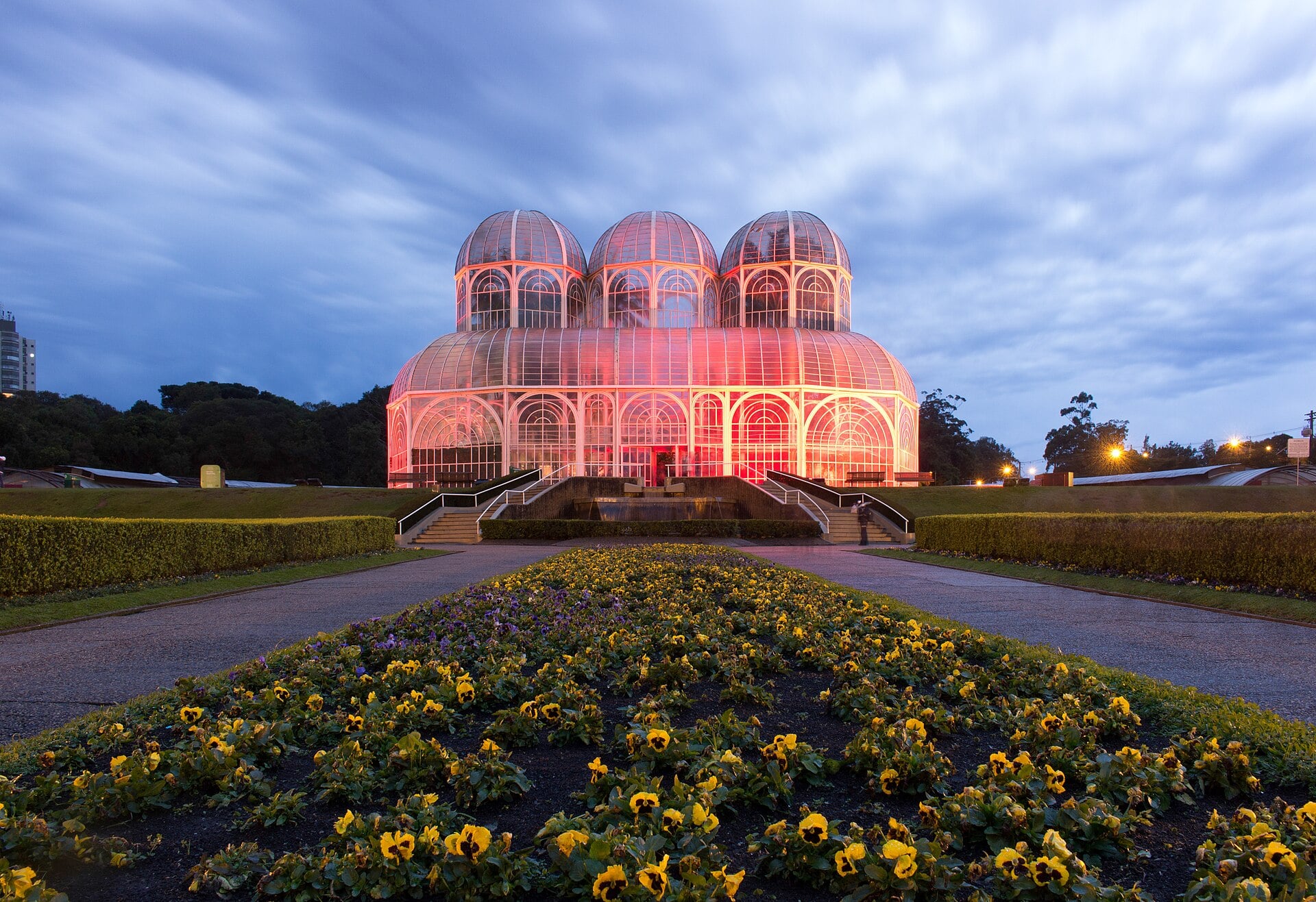 The glass and iron greenhouse of Curitiba's botanical garden lit at dusk, with formal flowerbeds of yellow and purple pansies in front.