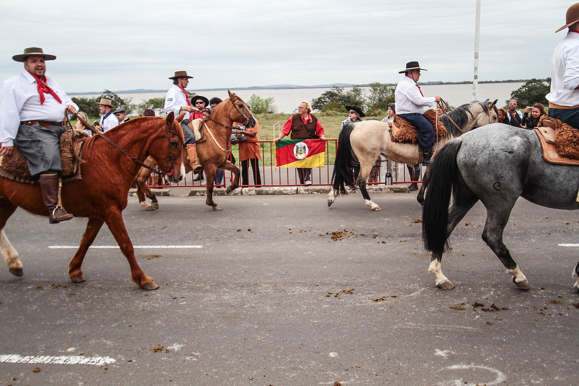 Riders in traditional gaúcho dress on horseback in Porto Alegre, with the Rio Grande do Sul state flag held up behind them.