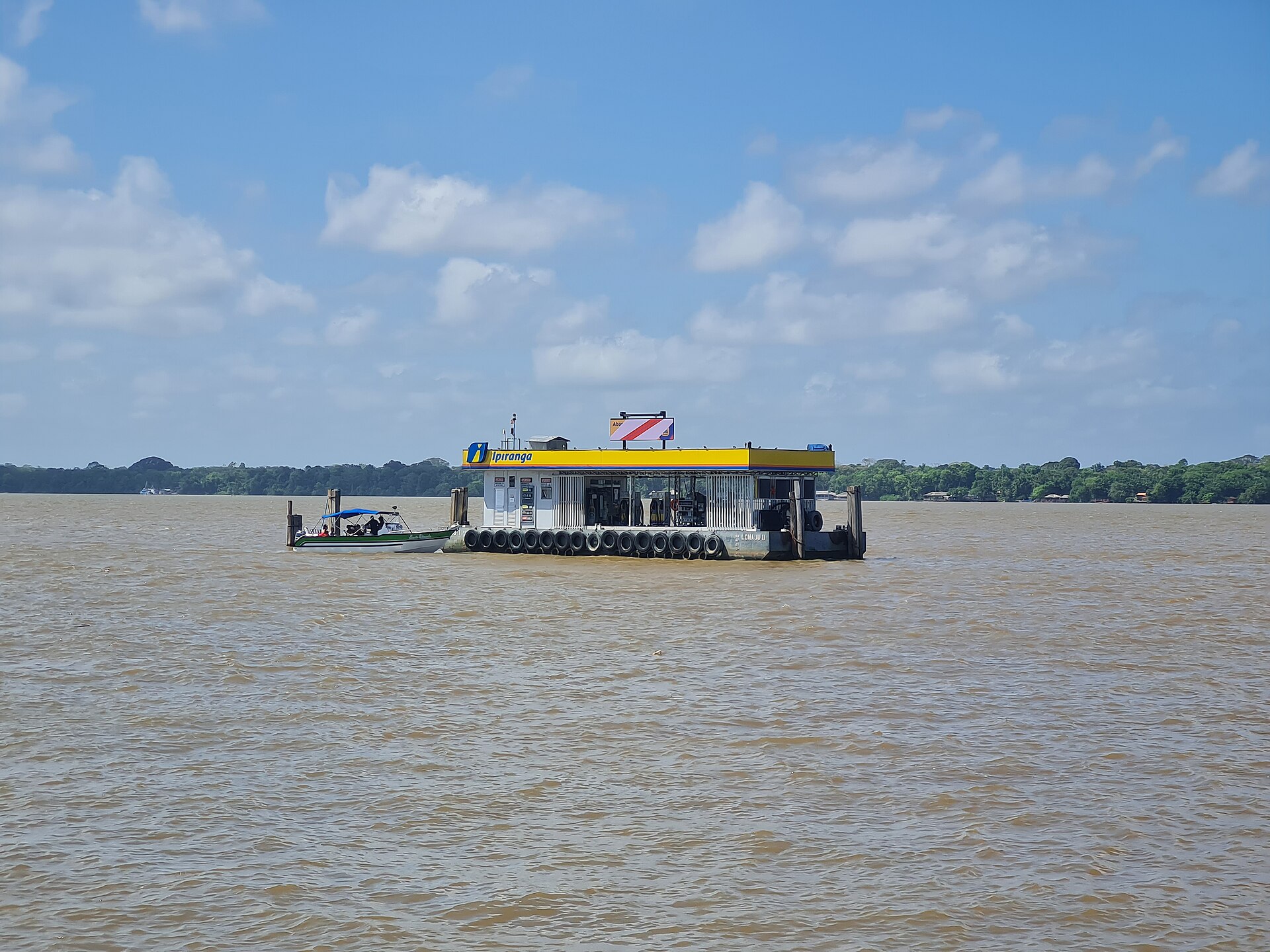 Floating Ipiranga fuel station moored on a wide brown river, with a small motorboat alongside and forest on the far bank