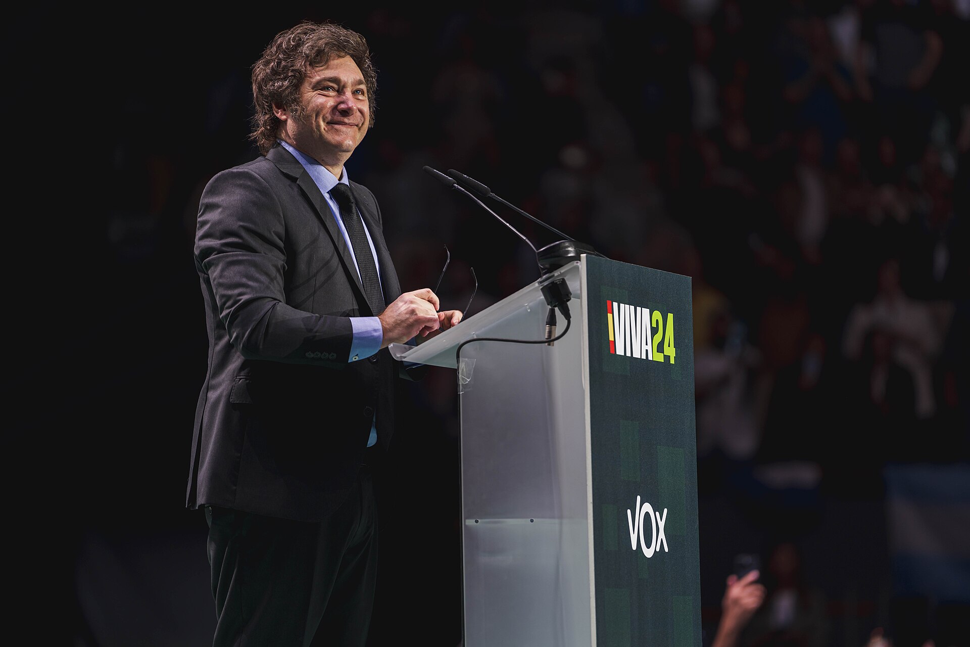 Javier Milei smiling at a lectern with microphones, holding his glasses, in front of a dark crowd