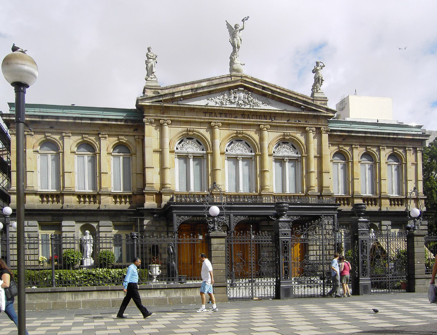 The neoclassical sandstone facade of the Teatro Nacional in San Jose, with statues on the pediment and pedestrians outside.