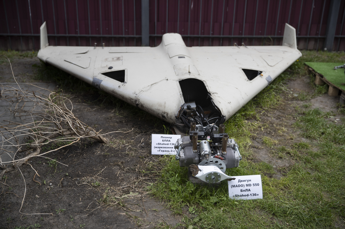 Wreckage of a white delta-wing attack drone and its separated engine, with Ukrainian-language labels, displayed on grass in front of a metal fence