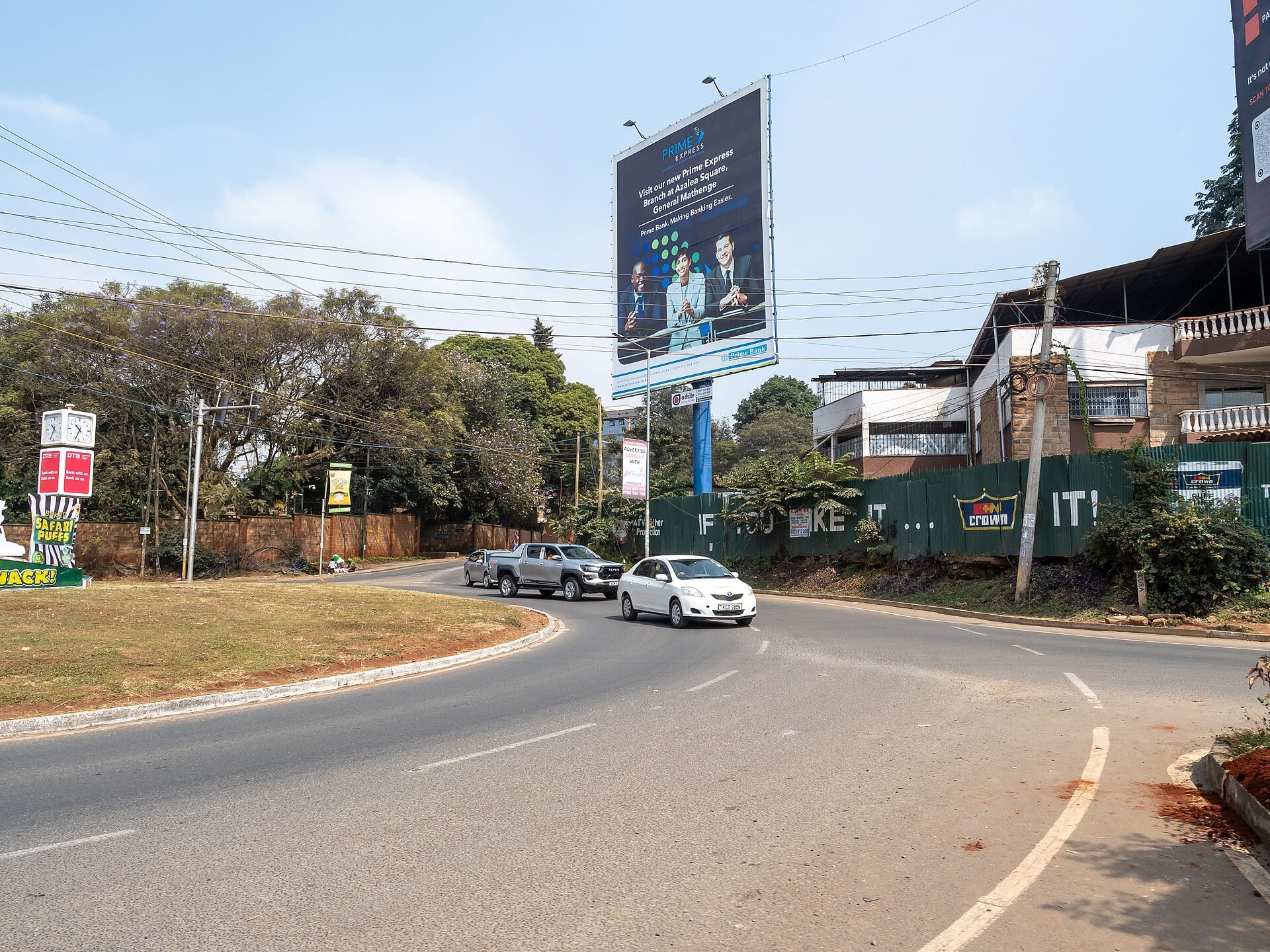 A roundabout in Nairobi with light traffic, a bank billboard, jacaranda trees and overhead power lines.