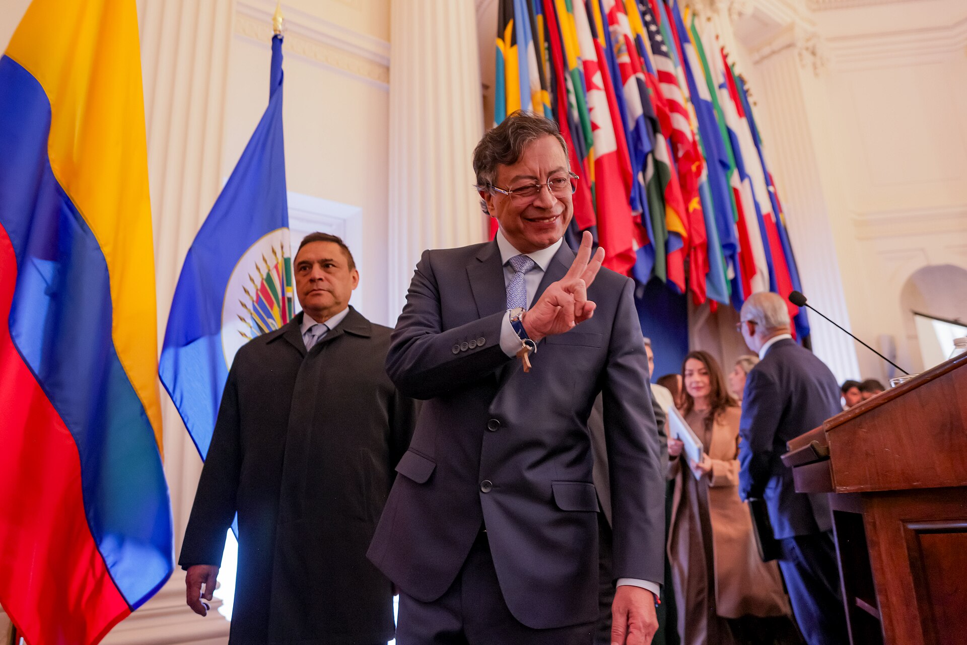 Gustavo Petro smiling and making a victory sign in a hall lined with national flags