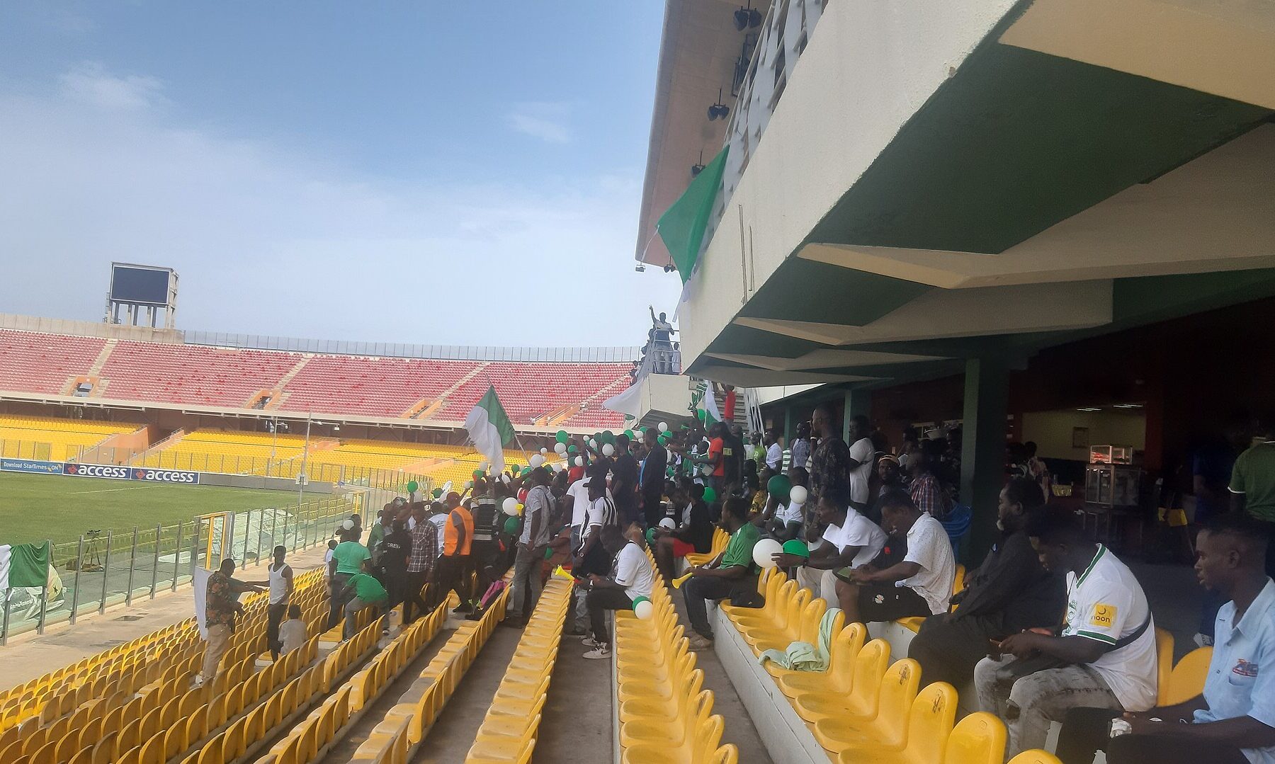 Supporters with Ghana flags and vuvuzelas in the yellow seats of the Accra Sports Stadium, the red stands empty behind them.