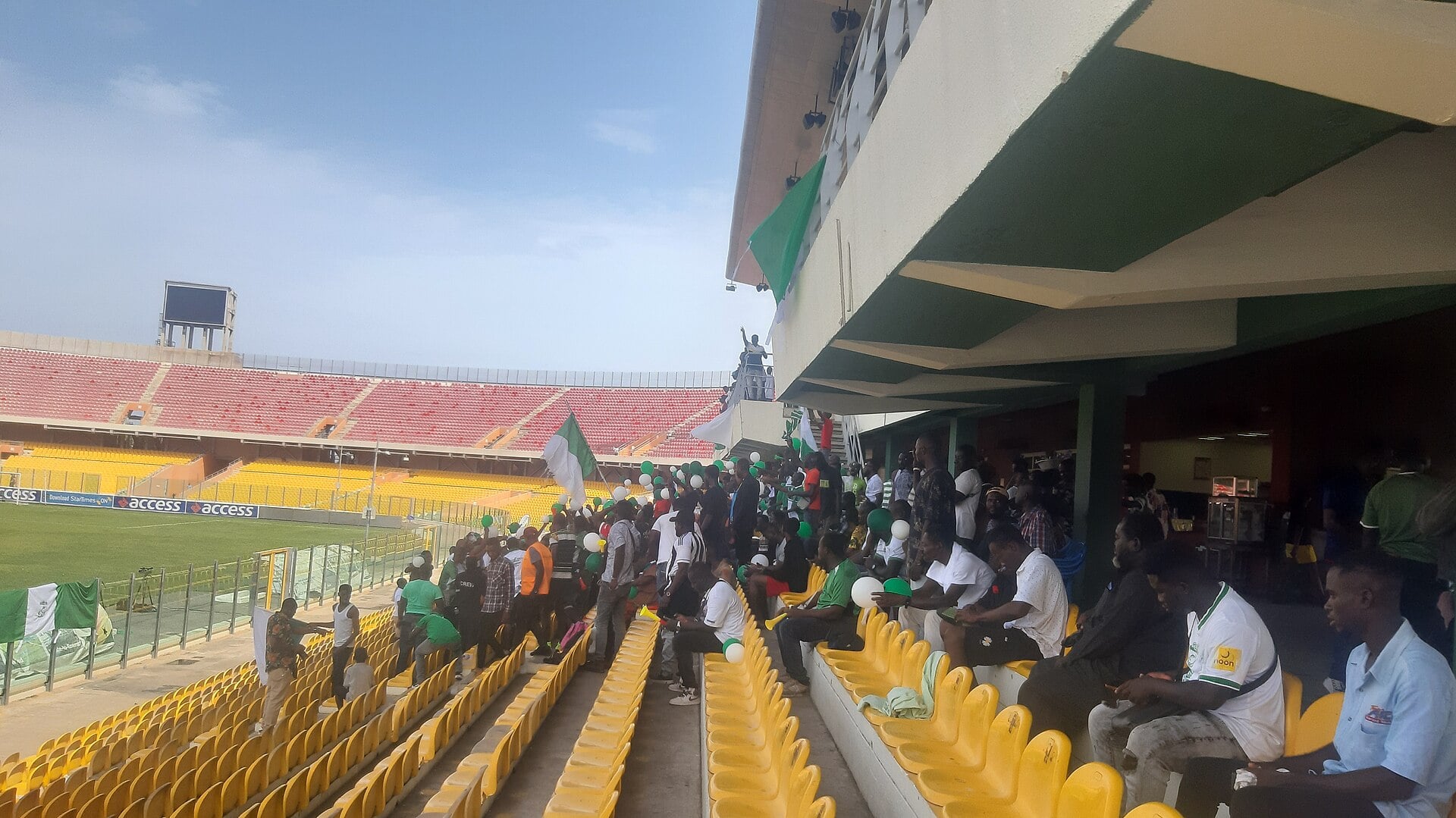 Supporters with Ghana flags and vuvuzelas in the yellow seats of the Accra Sports Stadium, the red stands empty behind them.