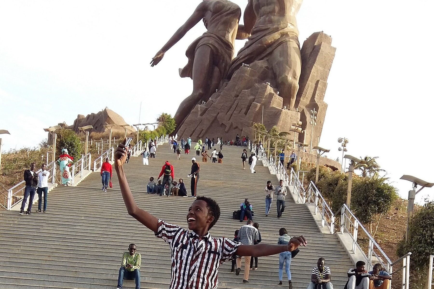 Young people on the steps below the African Renaissance Monument in Dakar, one lifted on another's shoulders with arms outstretched.