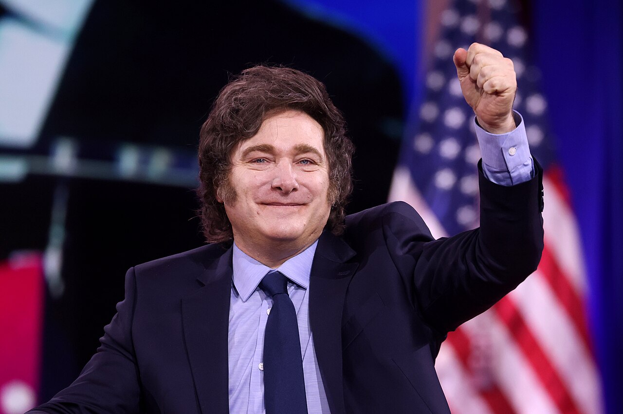 Milei poll: Argentina’s President Javier Milei smiles and raises his fist on stage at a conference, with a US flag behind him