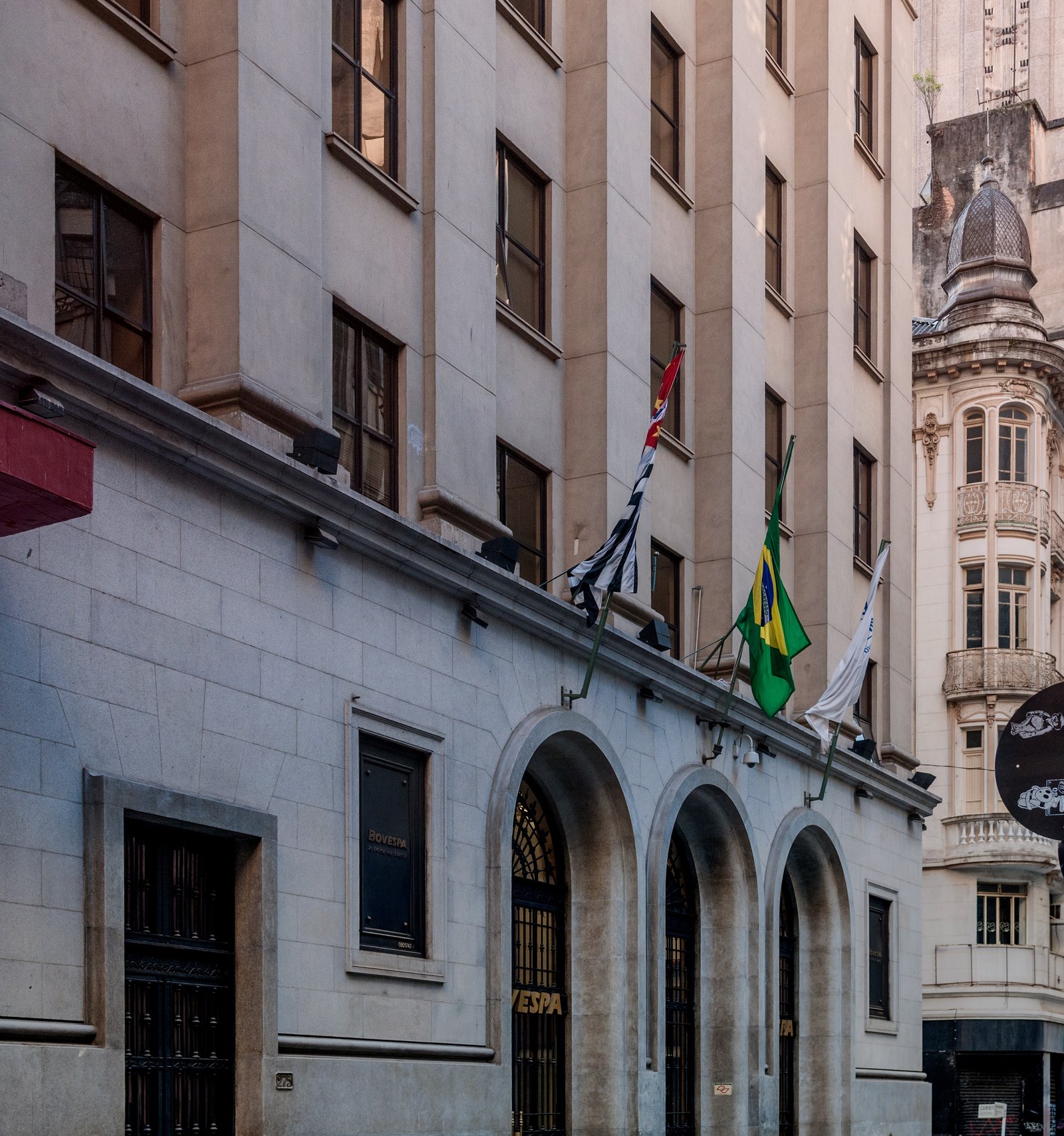 The ornate stone facade and arched entrance of the B3 stock exchange building in downtown São Paulo, with Brazilian and São Paulo state flags flying above the doorway