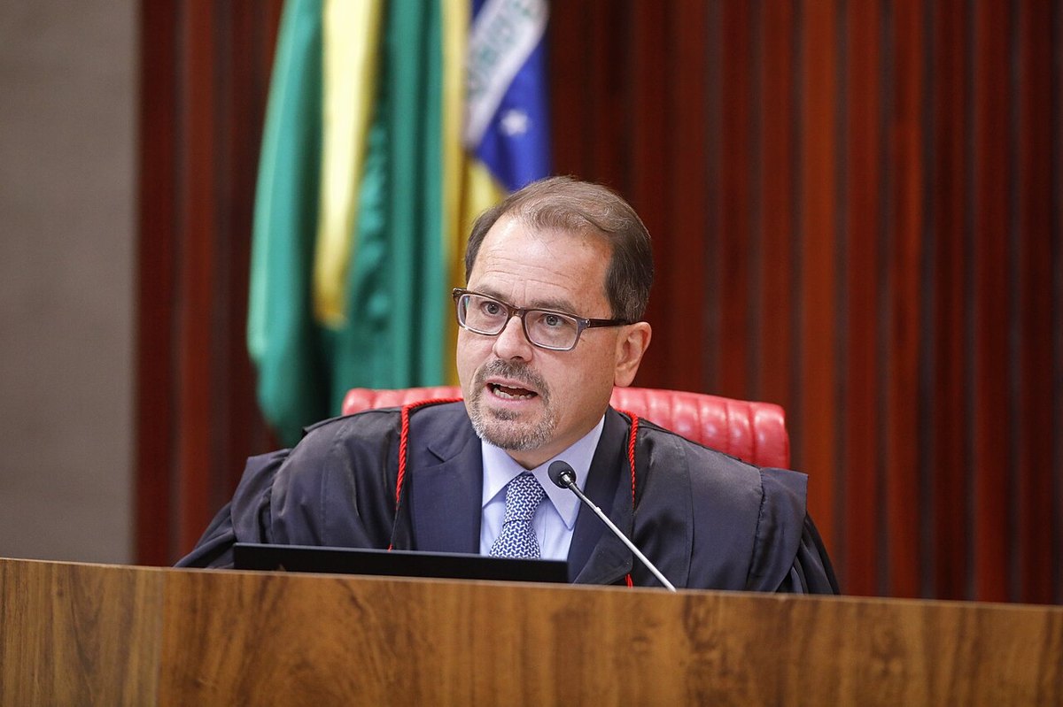A justice in black robes speaking into a microphone at the bench of Brazil's Superior Electoral Court, with flags behind