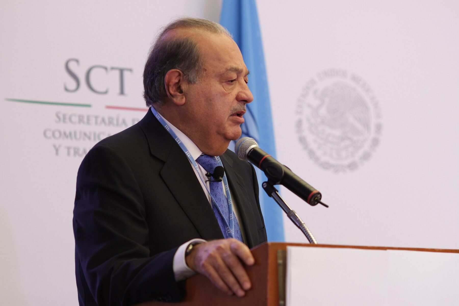 Carlos Slim speaks into a microphone at a podium during a press event, with a Mexican government seal in the background