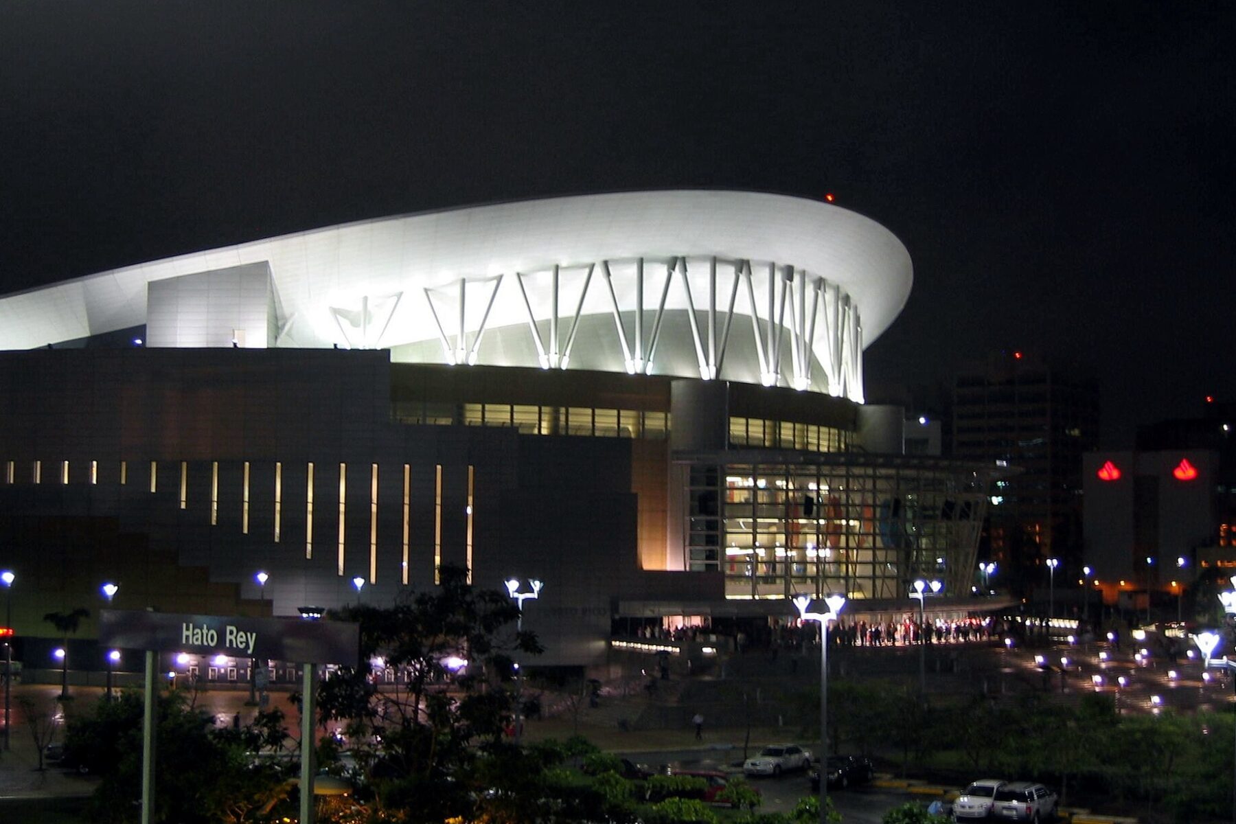 The Coliseo de Puerto Rico lit white at night, its curved roof above the entrance plaza, with a Hato Rey station sign in front.