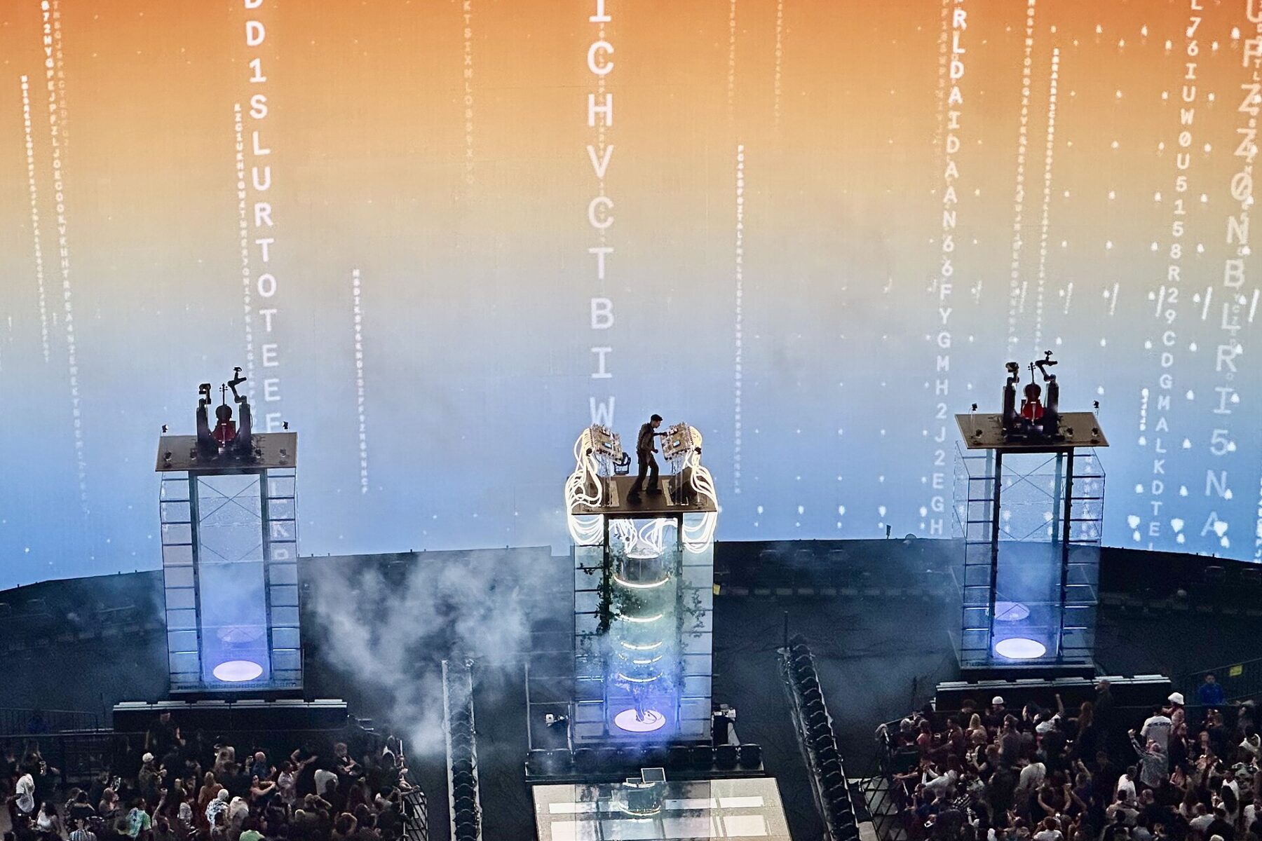 Anyma standing on a lit glass tower at the Sphere in Las Vegas, two towers with robotic arms holding cellos beside him, crowd below