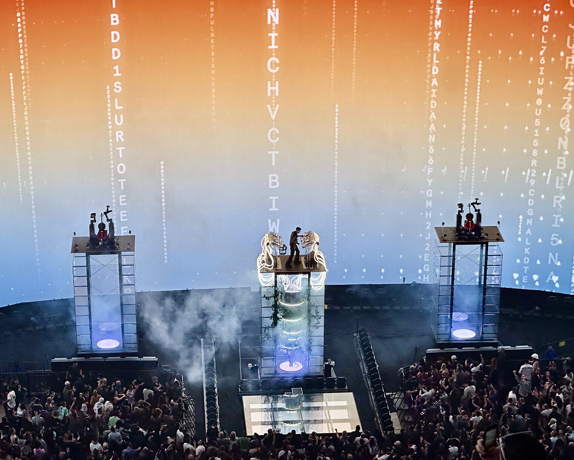 Anyma standing on a lit glass tower at the Sphere in Las Vegas, two towers with robotic arms holding string instruments beside him, crowd below