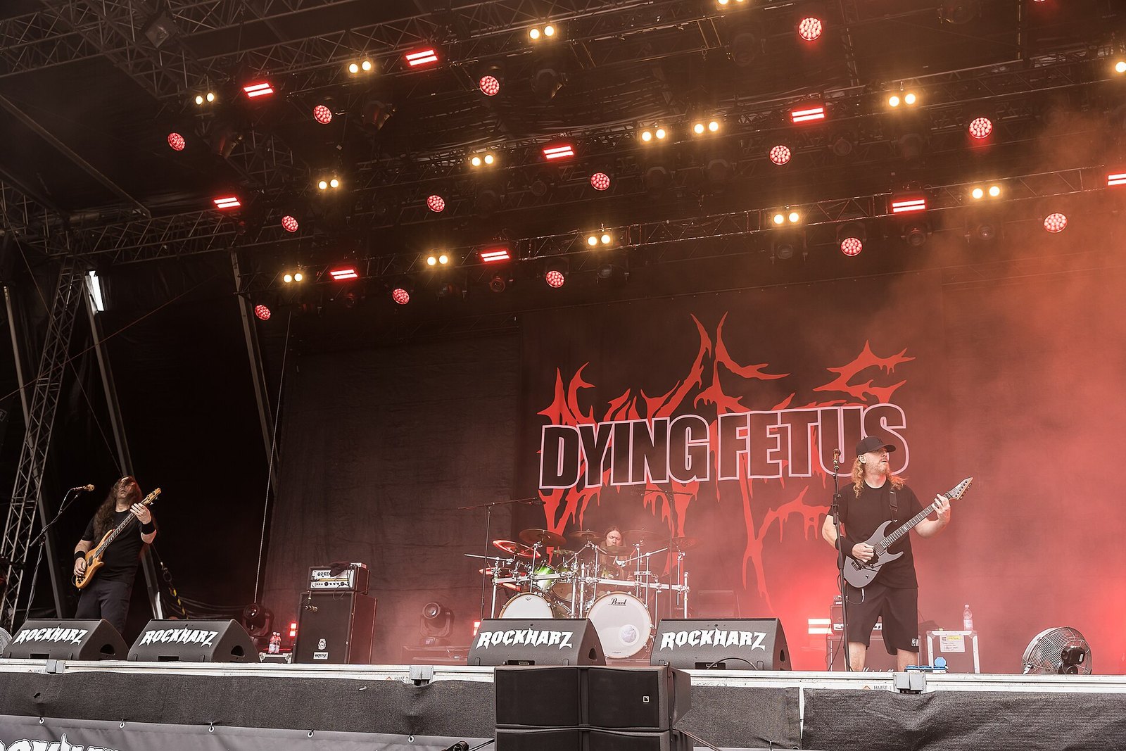 Dying Fetus on an outdoor festival stage: a long-haired bassist on the left, the drummer in the centre and a guitarist in a black cap on the right, in front of a black backdrop with the band's name and red logo