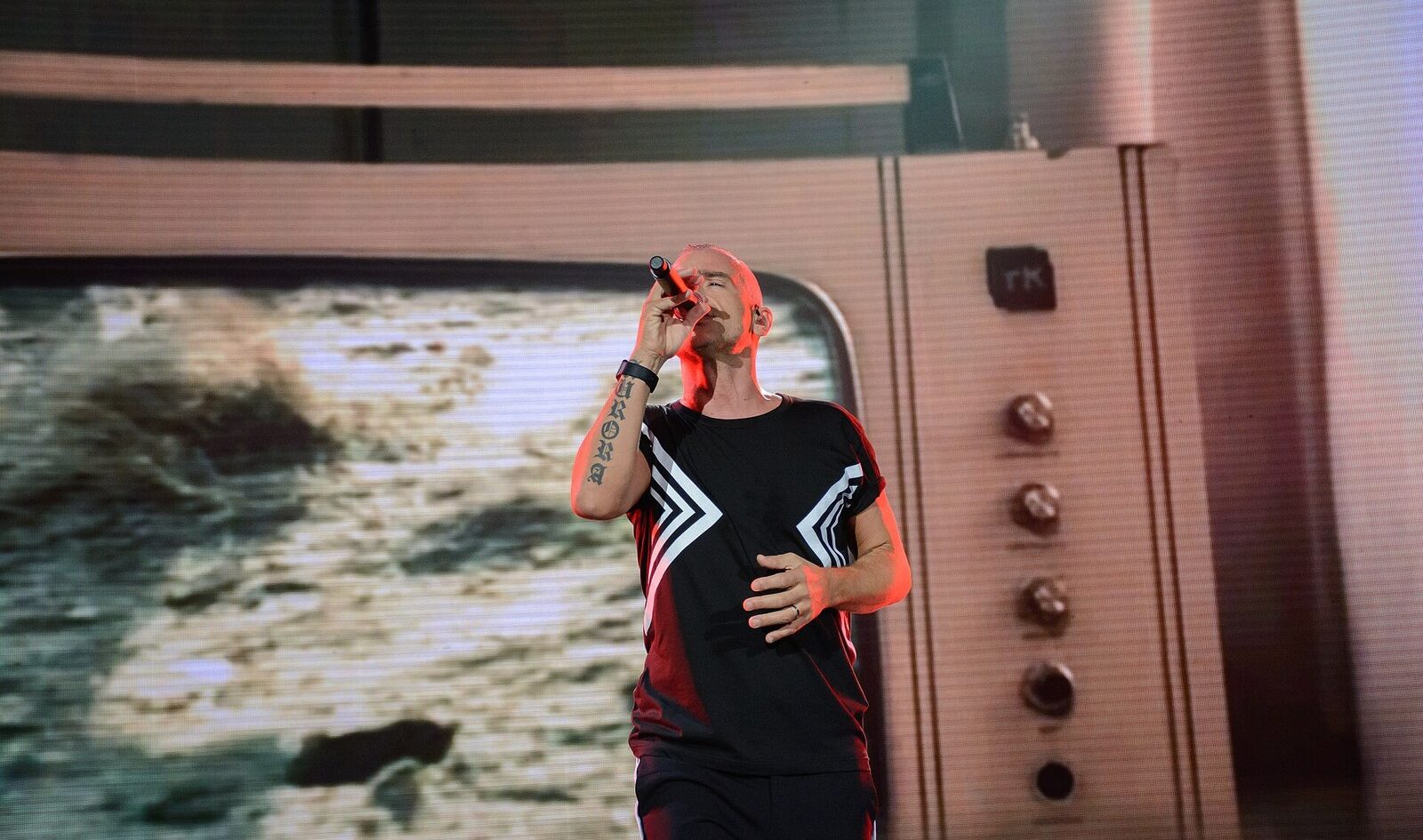 Eros Ramazzotti sings into a handheld microphone on stage, in a black T-shirt with white chevrons, a giant screen styled as a vintage television behind him