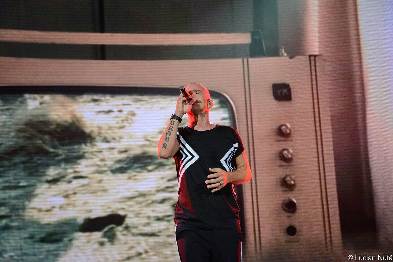 Eros Ramazzotti sings into a handheld microphone on stage, in a black T-shirt with white chevrons, a giant screen styled as a vintage television behind him