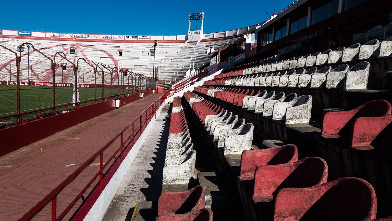 Empty red and white concrete seats along the side of Estadio Huracán in Buenos Aires, with the pitch behind a fence and the far stand under a clear blue sky