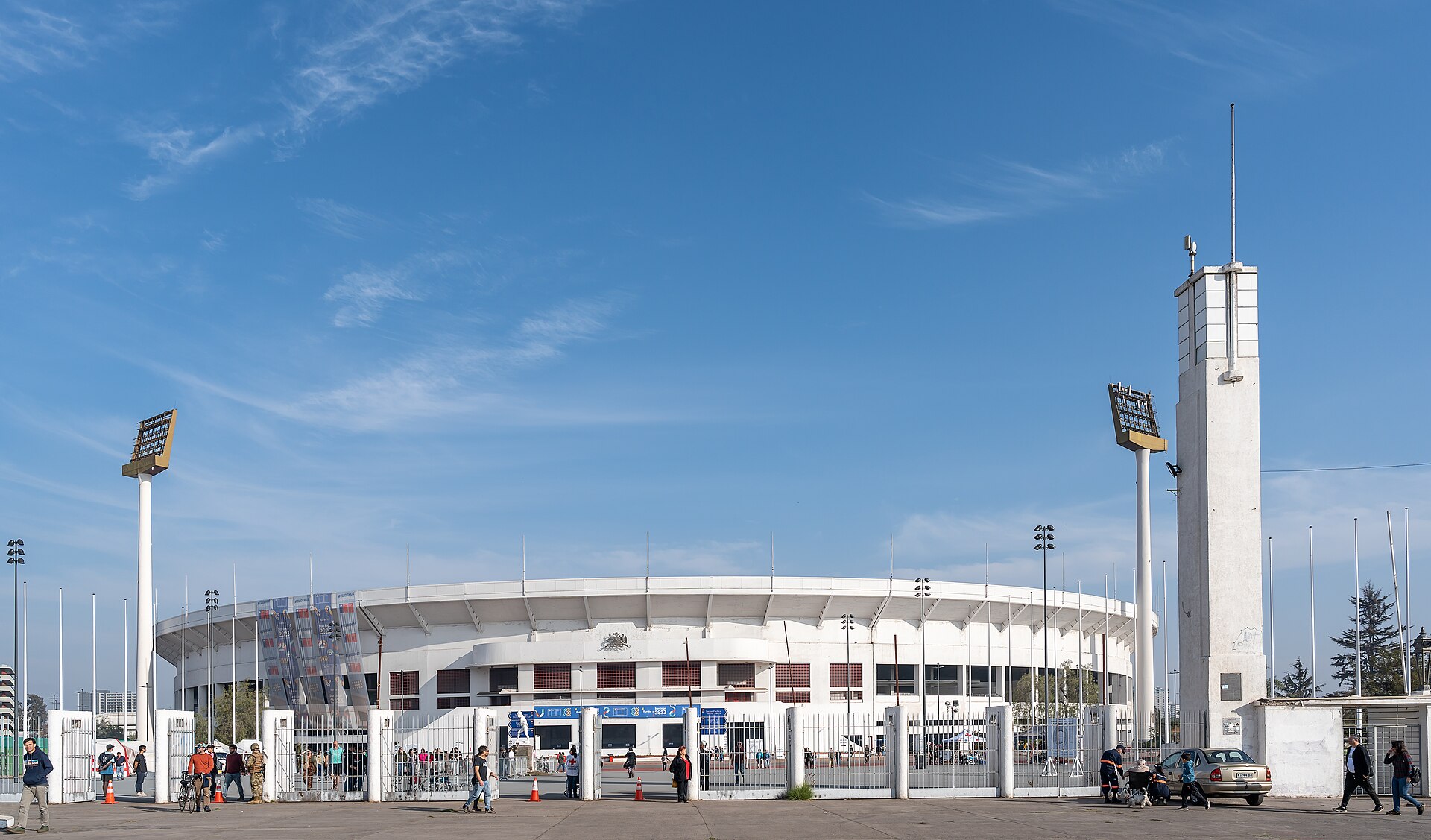 The white oval Estadio Nacional in Santiago behind its entrance gates under a blue sky, with floodlight towers, a tall white tower and people walking past