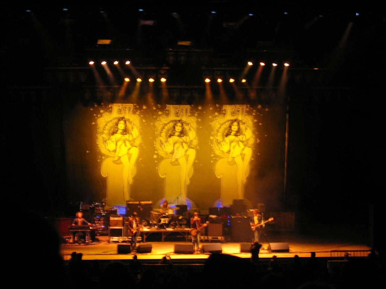 JET performing on a stage lit in yellow, five musicians in front of three banners with the band's logo and an illustrated woman, audience silhouettes in the foreground