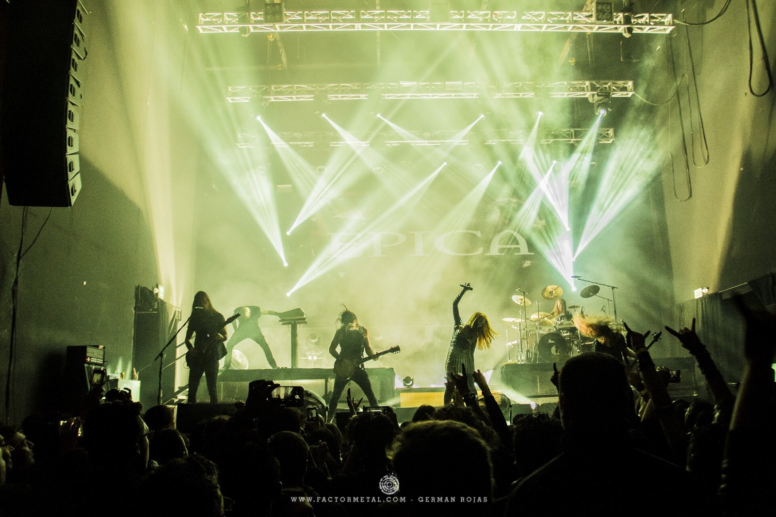 Band on a stage under white and green light beams, the name EPICA on the backdrop, silhouettes of fans with raised hands in front