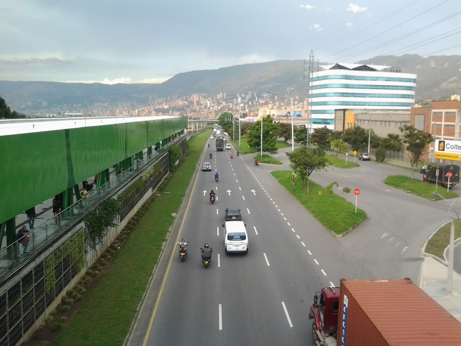 Motorbikes, a white van and a lorry on a multi-lane road beside a green elevated Metro walkway and a glass office block, with high-rises and mountains behind