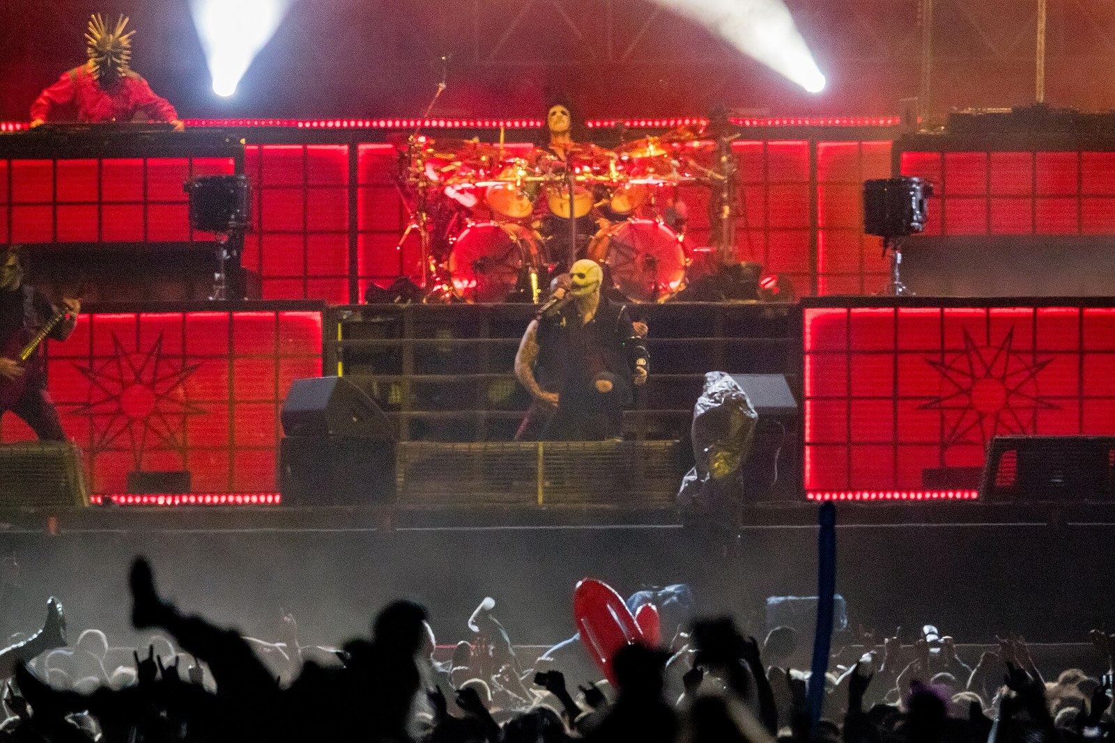 Slipknot's masked singer holds a microphone at the front of a red-lit stage, a masked drummer behind him and fans raising their hands in the foreground