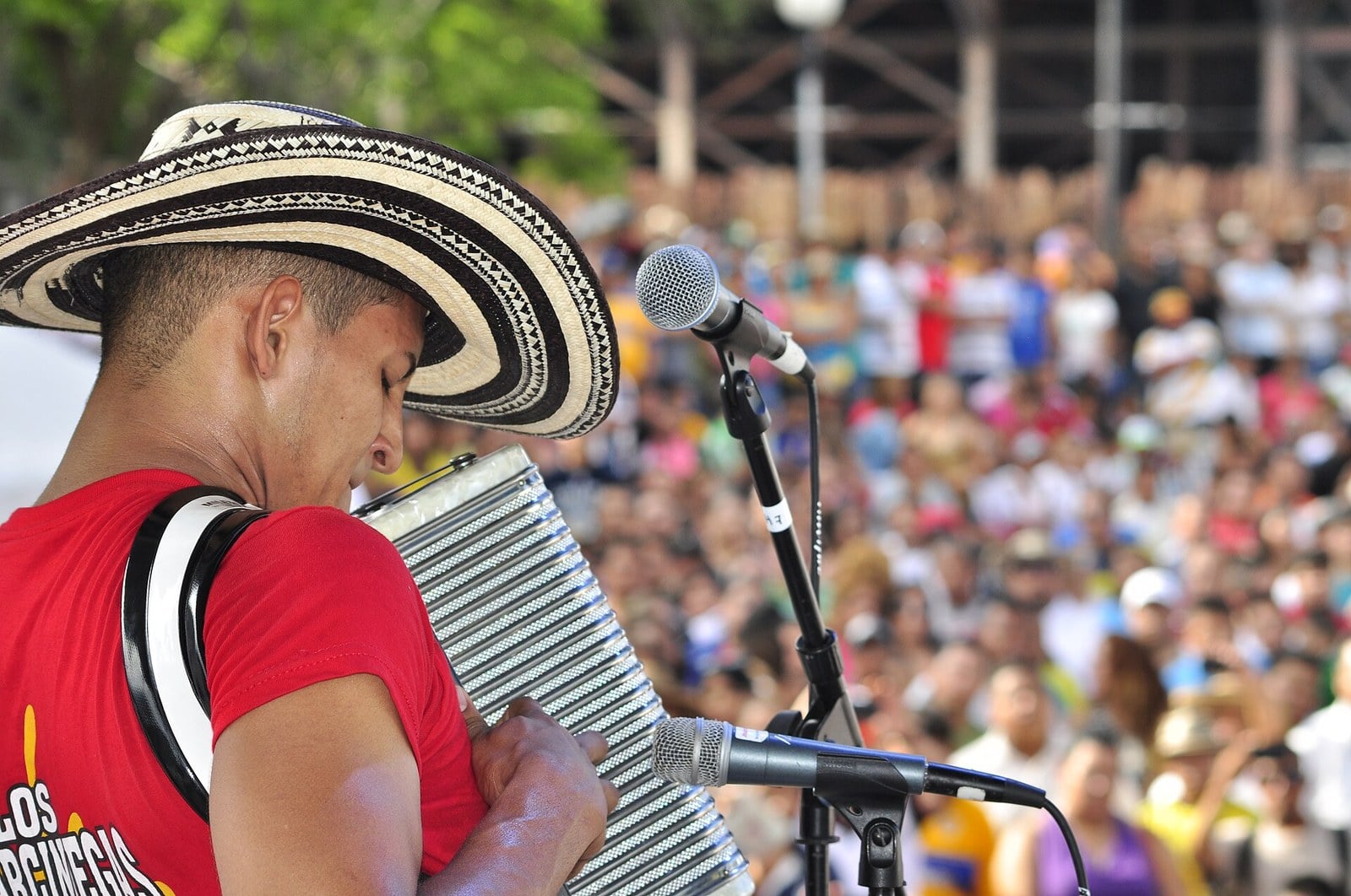 A young accordionist in a black-and-white woven hat and a red T-shirt plays a button accordion at a microphone before a large outdoor crowd