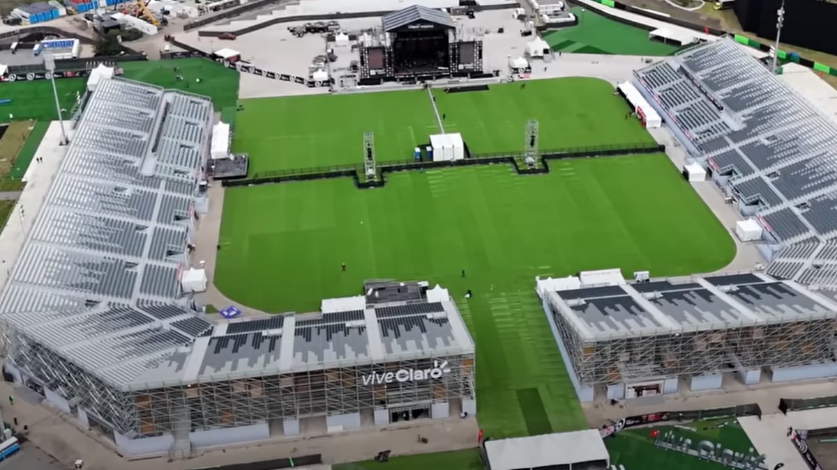 an empty grass field ringed by grey modular grandstands, with a covered stage at the far end