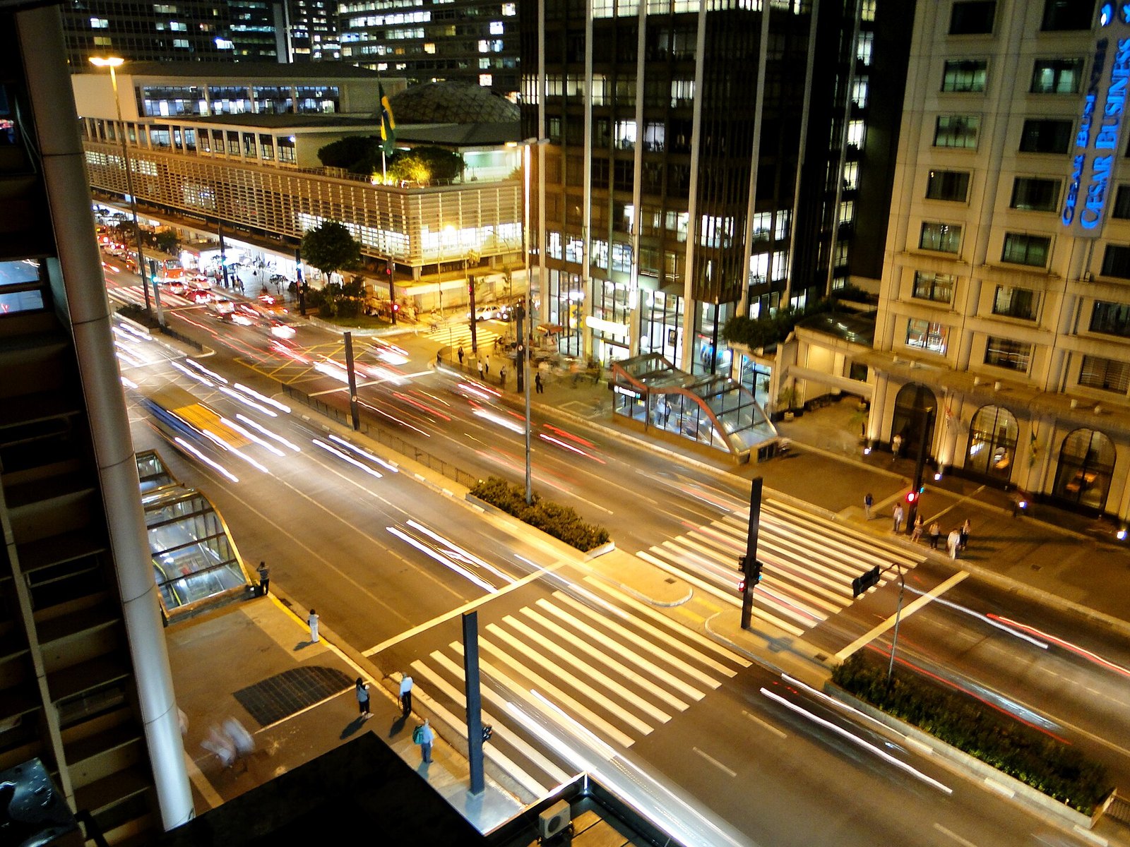 The Conjunto Nacional building lit up at night on Avenida Paulista, with light trails from traffic, pedestrian crossings and a glass-roofed metro entrance