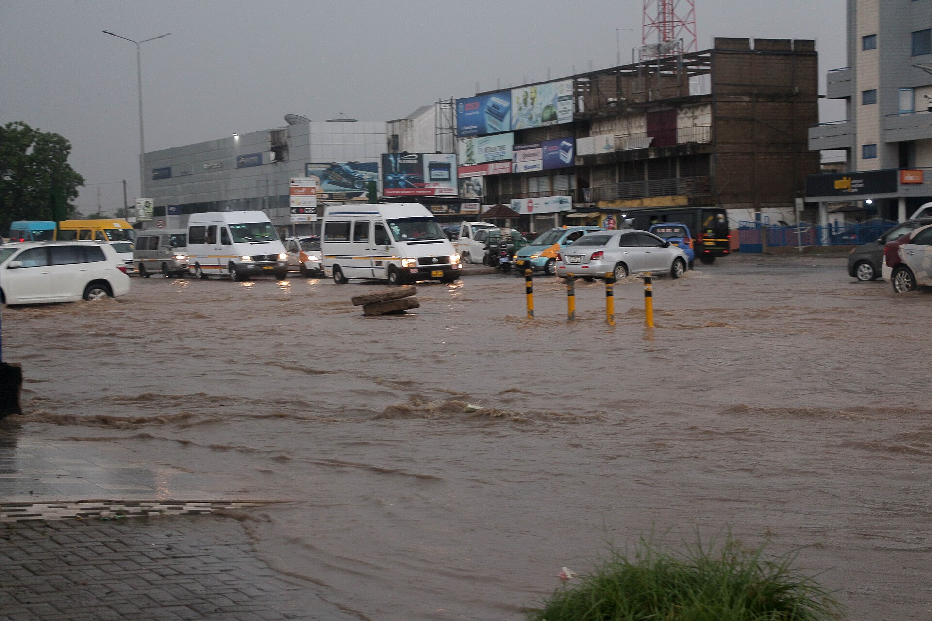 Floodwater covering a busy street in urban Ghana during a rainstorm, with minibuses and cars wading through