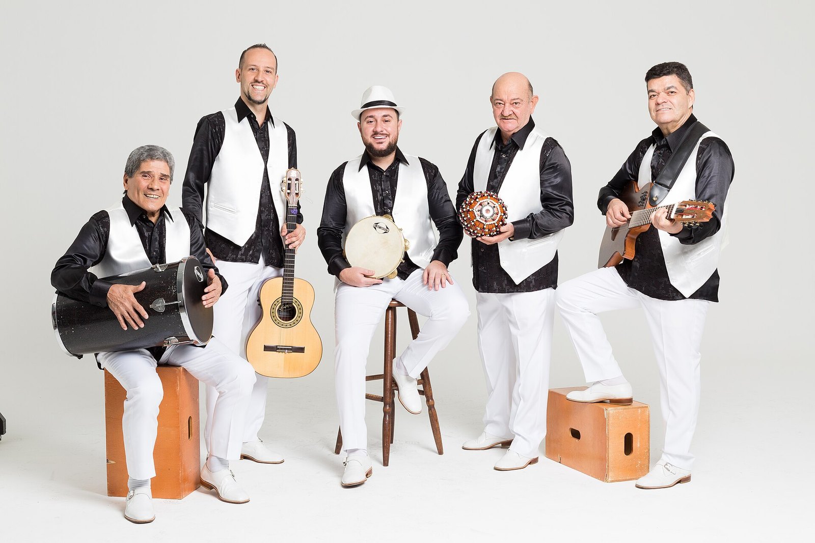 The five members of Demônios da Garoa in black shirts and white waistcoats, posing with a cavaquinho, guitar, tambourine, surdo drum and shaker